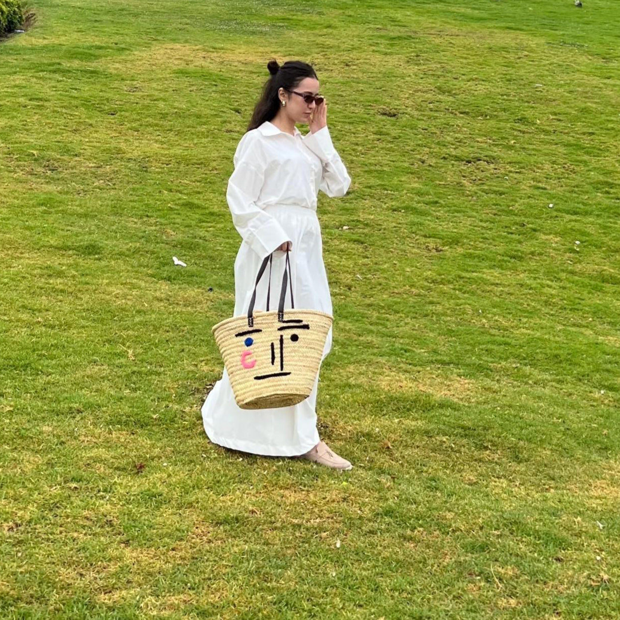 Woman in a white dress holding a woven bag with a face design on a grassy field.