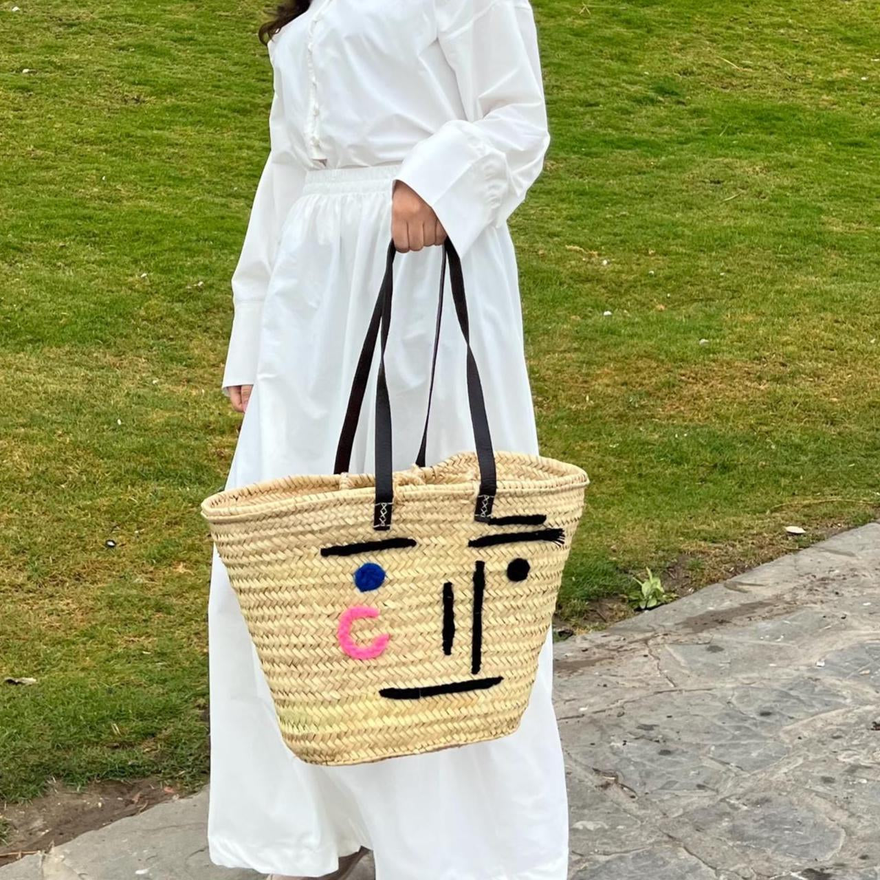 Woman in a white dress holding a woven bag with facial expressions on a grassy field.